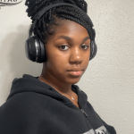 Young woman with braided hair wearing headphones and a black hoodie, looking at the camera against a plain wall.
