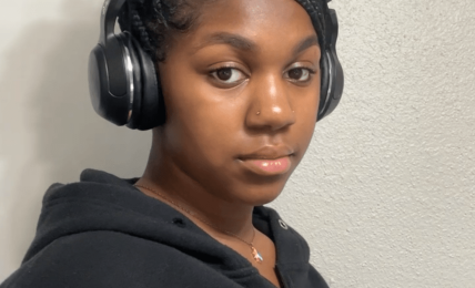 Young woman with braided hair wearing headphones and a black hoodie, looking at the camera against a plain wall.