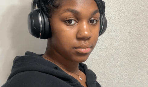 Young woman with braided hair wearing headphones and a black hoodie, looking at the camera against a plain wall.