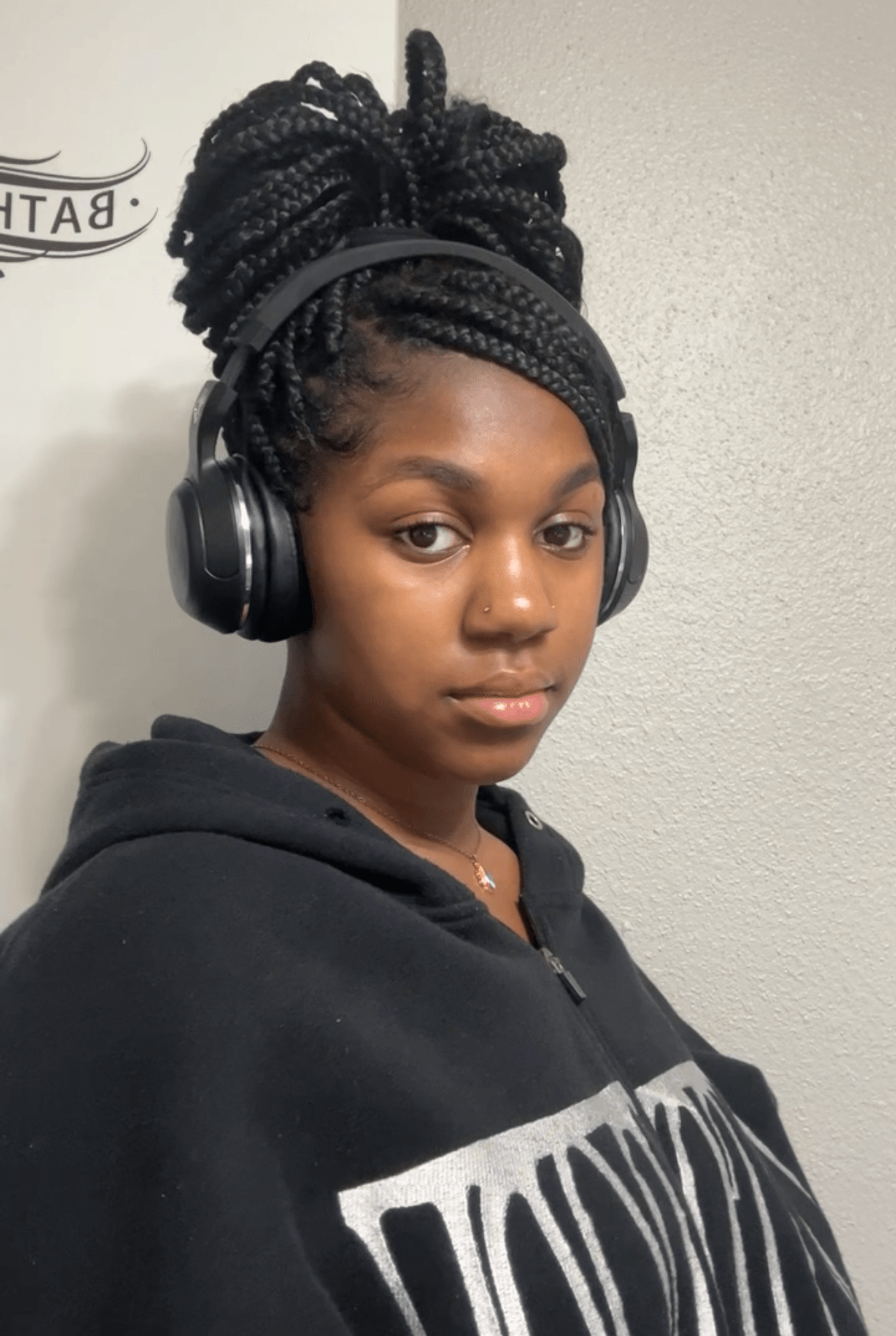 Young woman with braided hair wearing headphones and a black hoodie, looking at the camera against a plain wall.