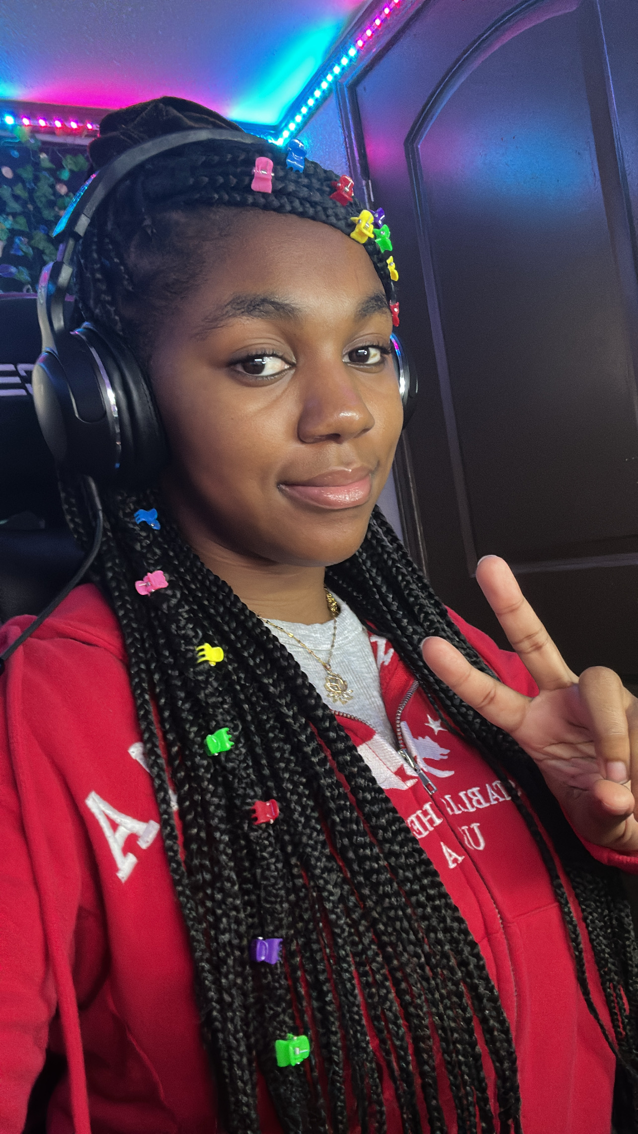 Young woman with braided hair, wearing headphones, making a peace sign, in front of colorful lights and a dark background.