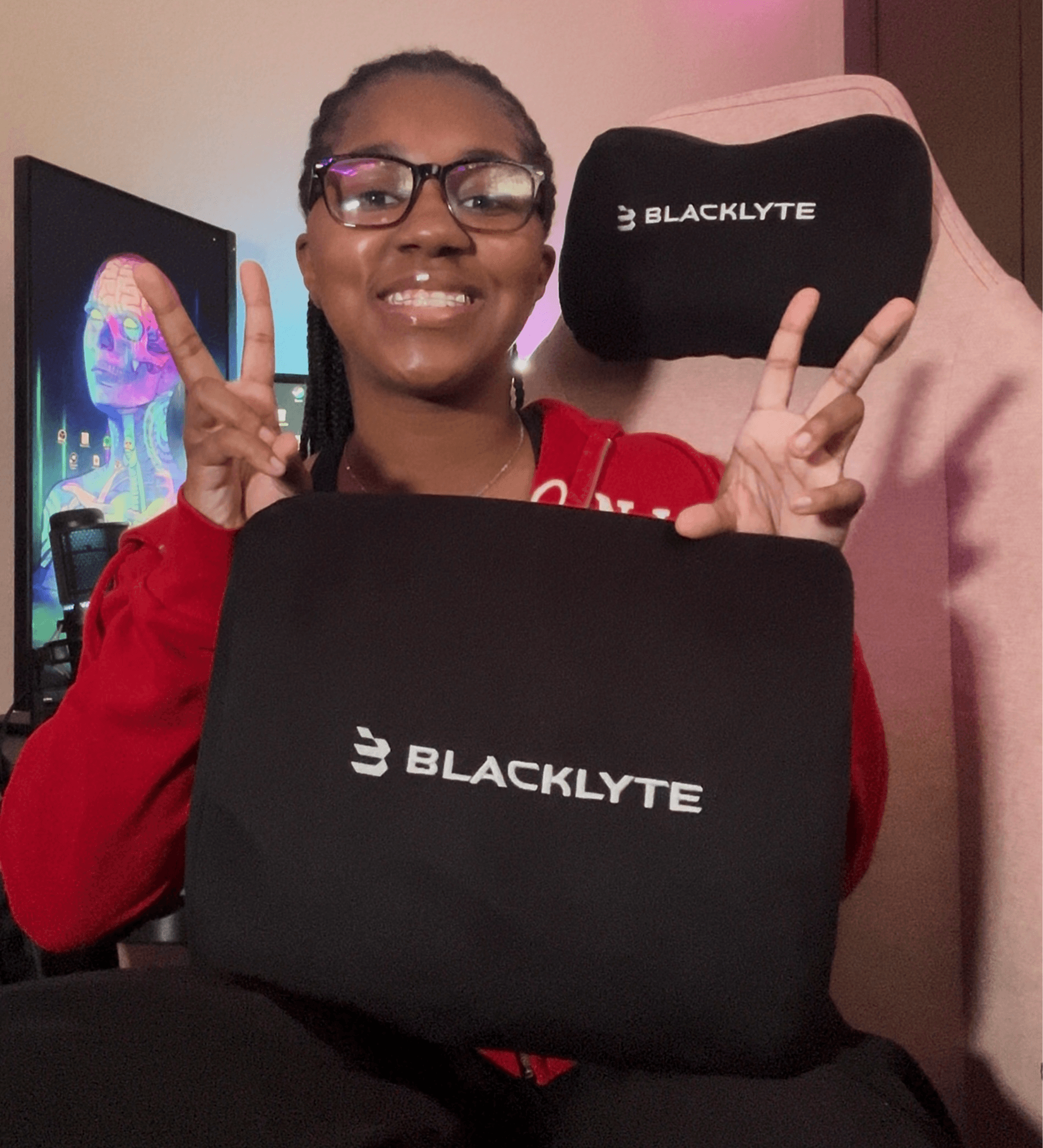 Person smiling with glasses, holding BlackKYTE branded items, making peace signs, in a room with a monitor in the background.