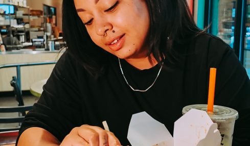 Woman with black hair sitting at a table, dipping food into sauce, in a cafe setting with colorful posters and large windows.