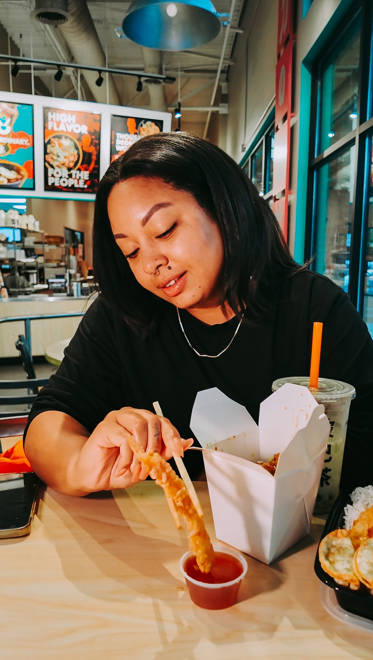 Woman with black hair sitting at a table, dipping food into sauce, in a cafe setting with colorful posters and large windows.