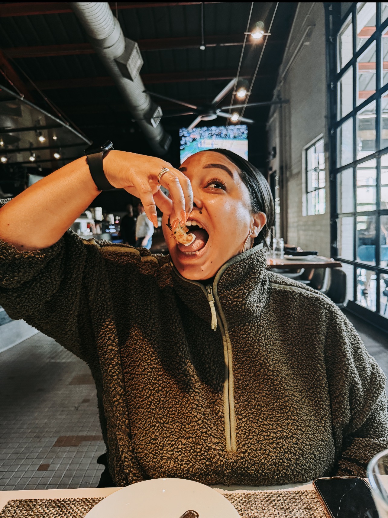 Person in a dark sweater eating food with an open mouth inside a modern restaurant with large windows.