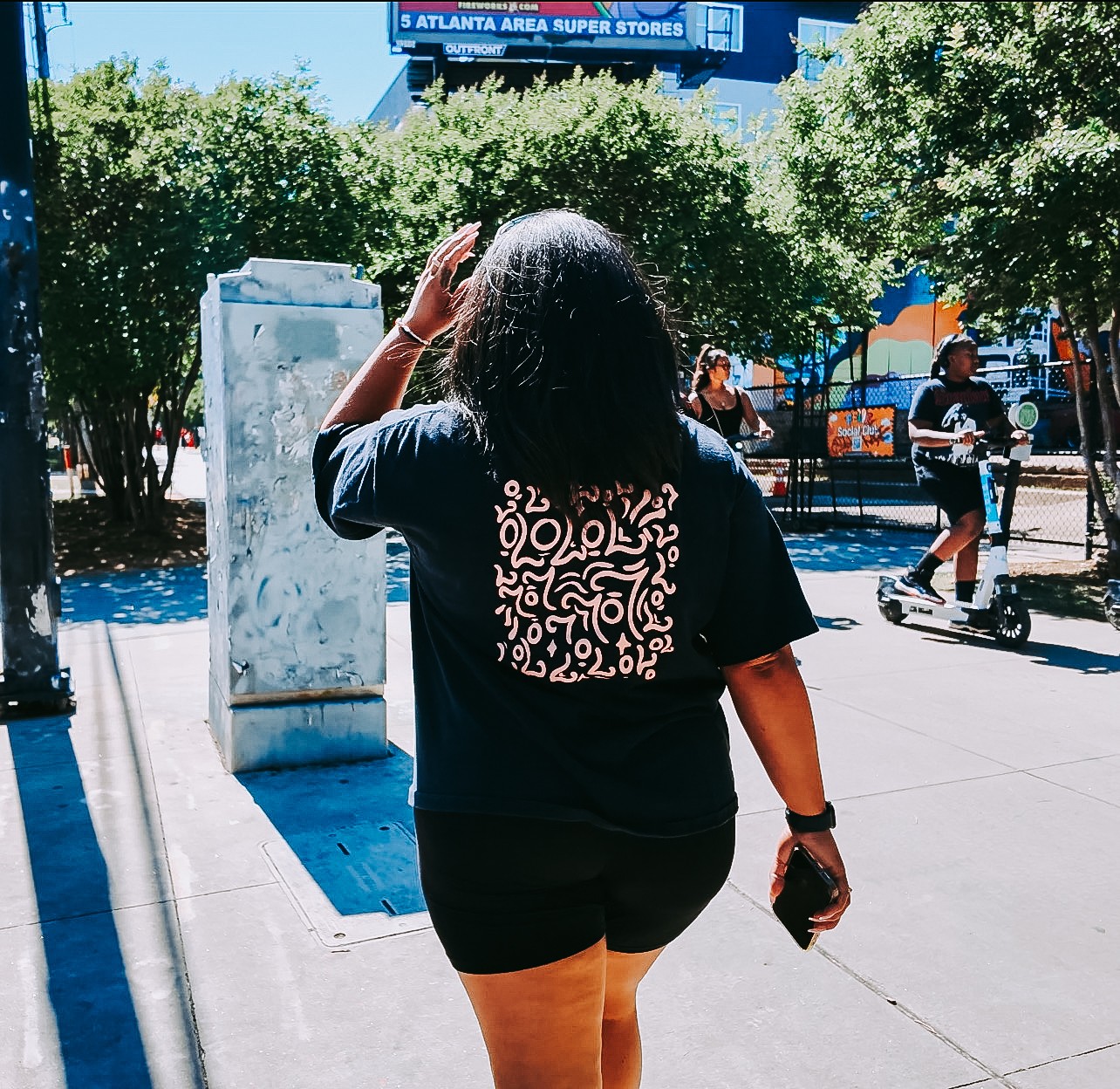 Person walking on sidewalk with trees and people in background, holding a phone, sunny day.