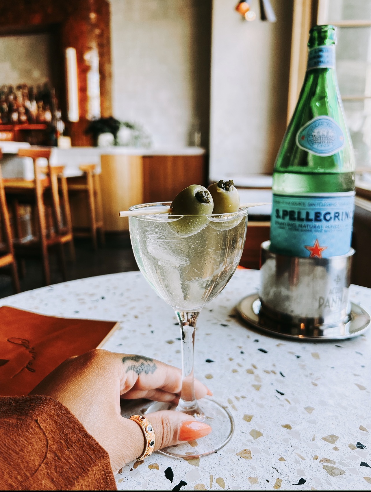 Hand holding a glass with green olives, on a speckled table in a restaurant setting.
