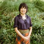 Young girl with short dark hair smiling outdoors, wearing a dark blouse and plaid skirt, surrounded by greenery.