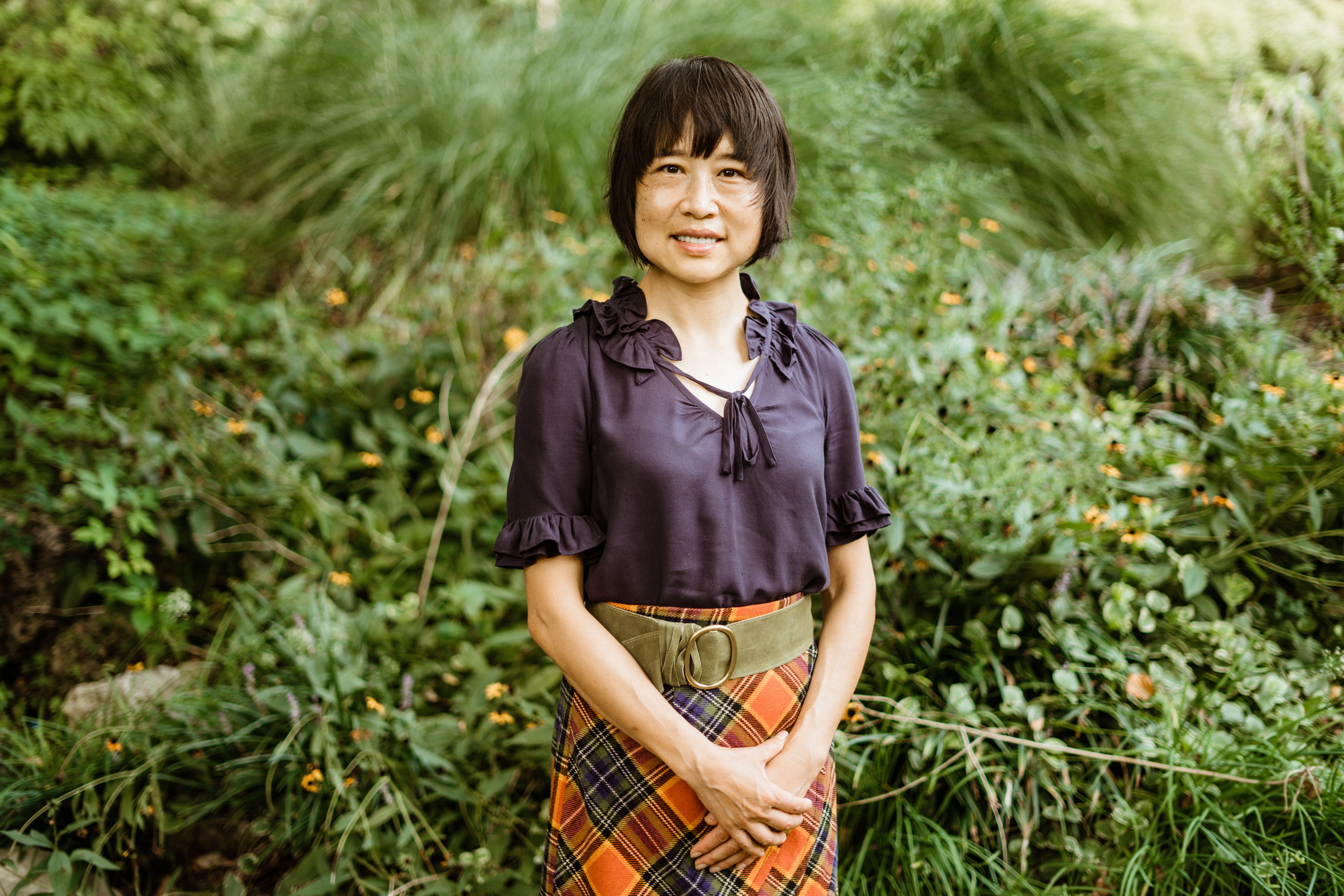Young girl with short dark hair smiling outdoors, wearing a dark blouse and plaid skirt, surrounded by greenery.
