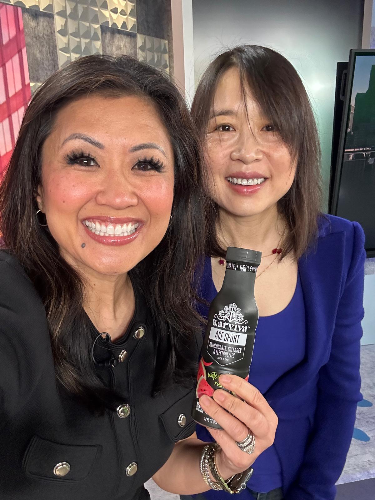 Two women smiling, one holding a microphone, in an indoor setting with a screen in the background.