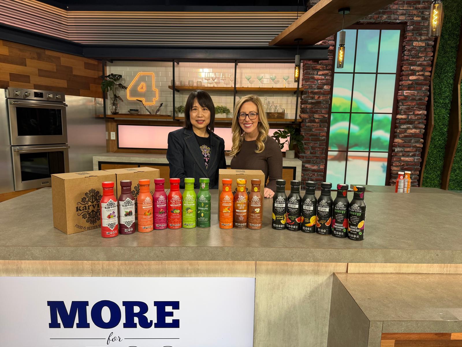 Two women stand behind a counter with colorful bottles of beverages, in a modern kitchen studio setting.