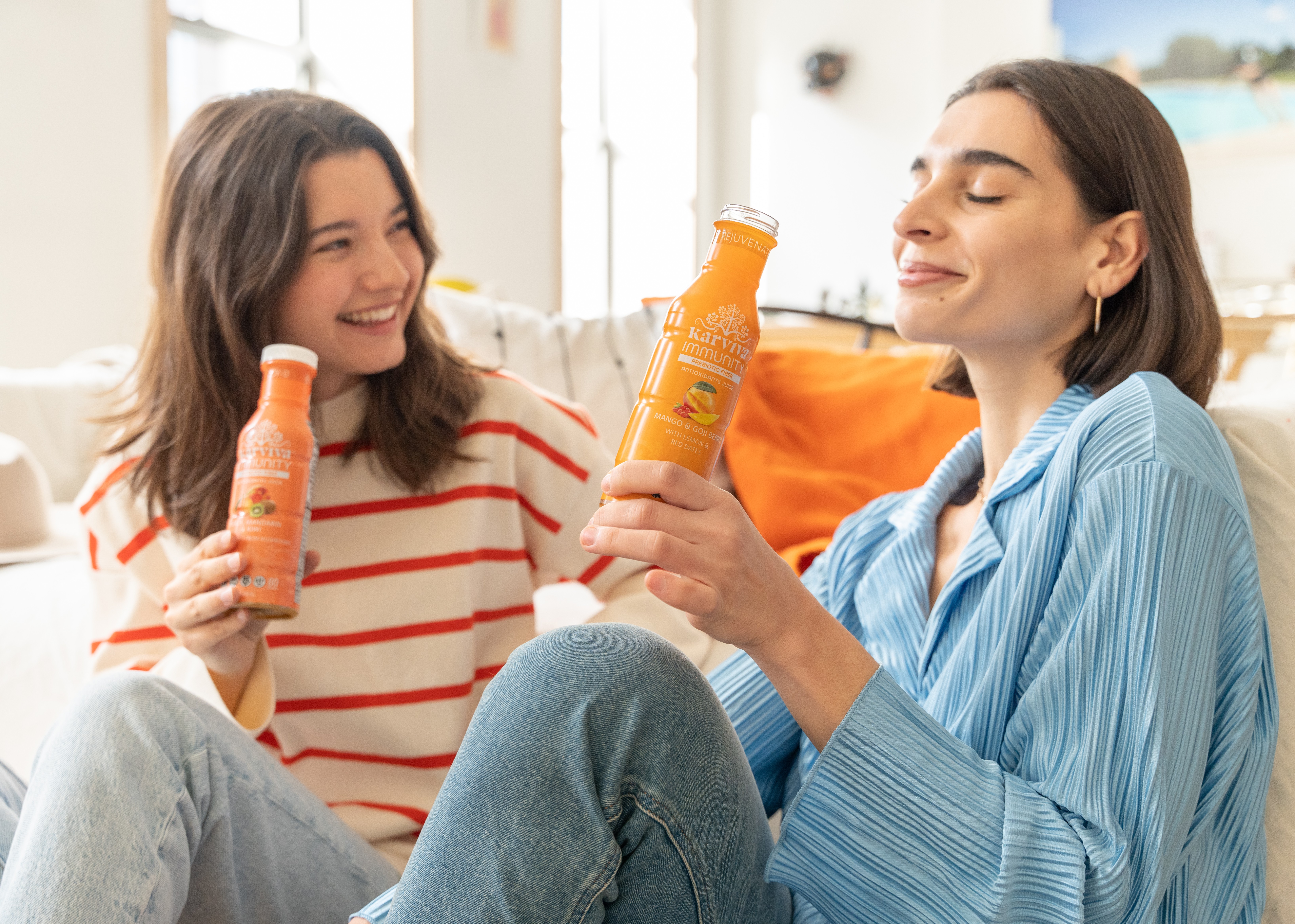 Two women sitting on a couch, smiling, holding bottles of orange-colored drinks, in a bright room with large windows.