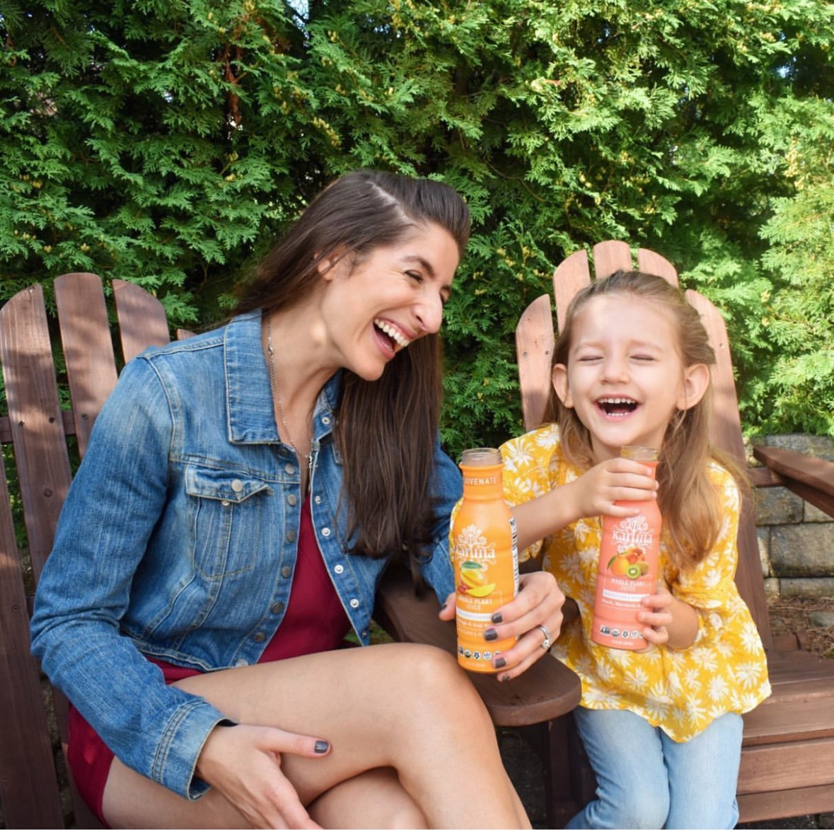 Woman and girl laughing outdoors, holding bottles, sitting on wooden chairs with green trees behind them.