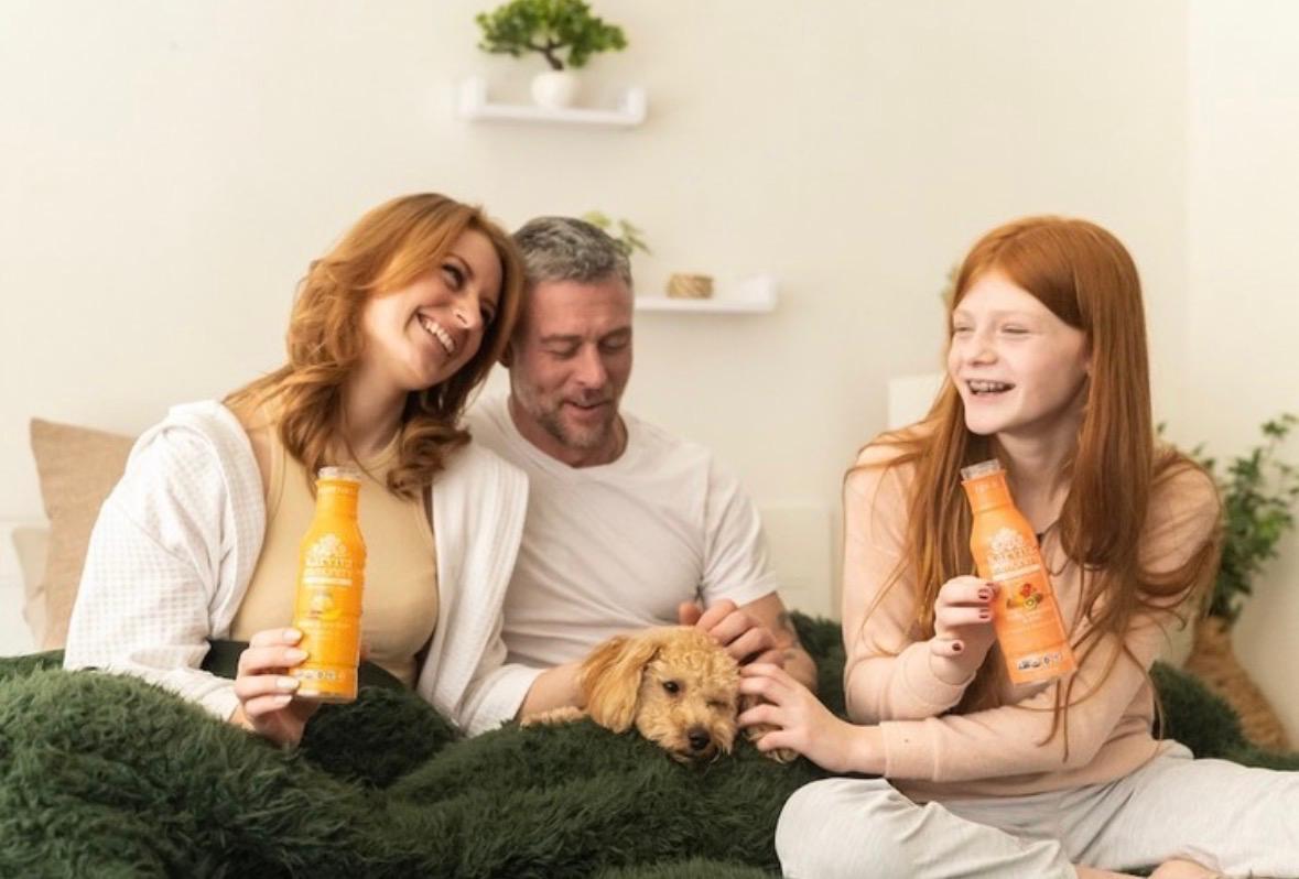 Three people and a dog on a bed, holding orange bottles, smiling, in a bright room with plants and shelves.