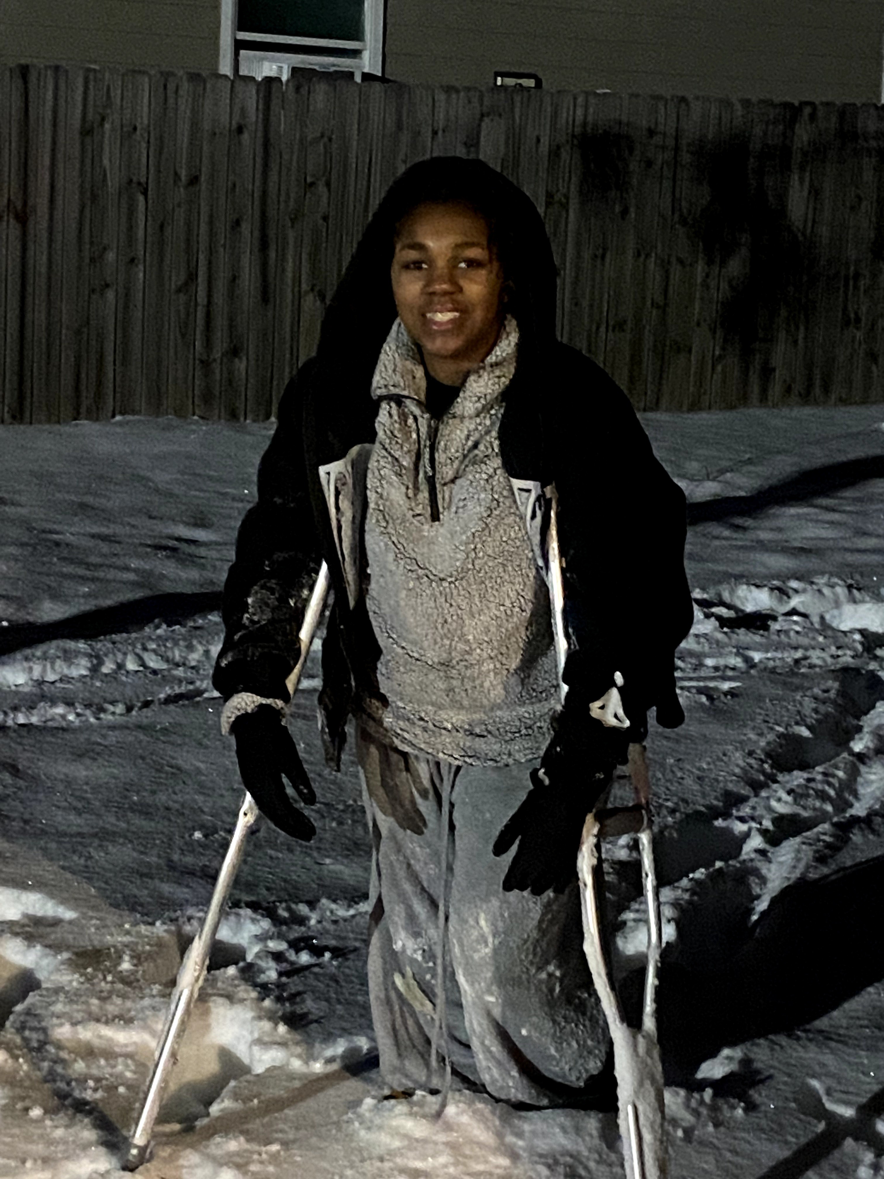 Person standing outdoors in snow at night, smiling, wearing a hoodie, jacket, and gloves, holding ski poles.
