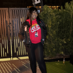 Person standing outdoors at night, smiling, wearing a red shirt, black jacket, black pants, and sneakers, near a gate and greenery.