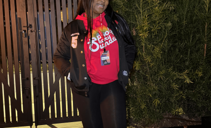 Person standing outdoors at night, smiling, wearing a red shirt, black jacket, black pants, and sneakers, near a gate and greenery.