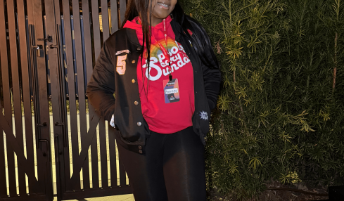 Person standing outdoors at night, smiling, wearing a red shirt, black jacket, black pants, and sneakers, near a gate and greenery.