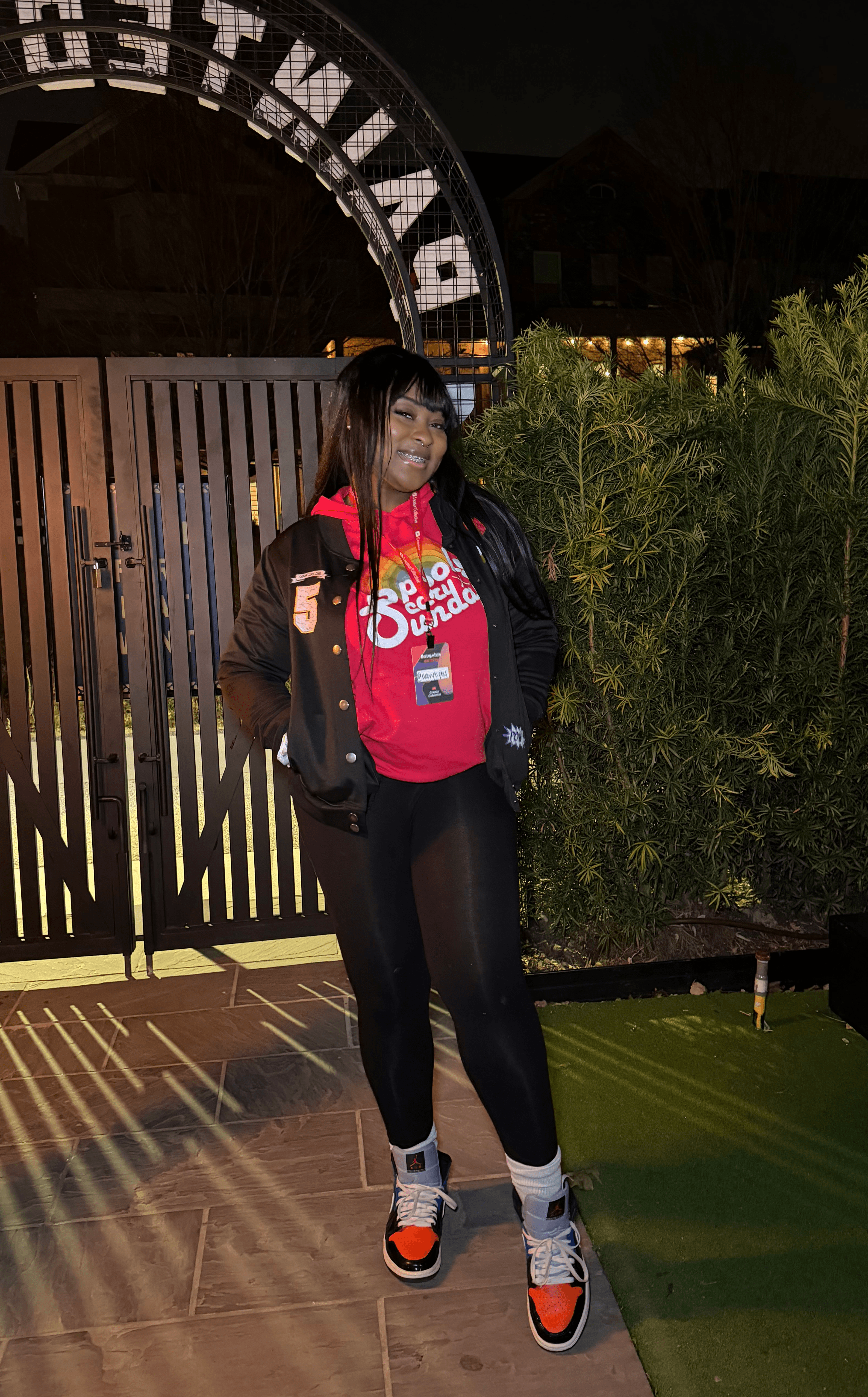 Person standing outdoors at night, smiling, wearing a red shirt, black jacket, black pants, and sneakers, near a gate and greenery.