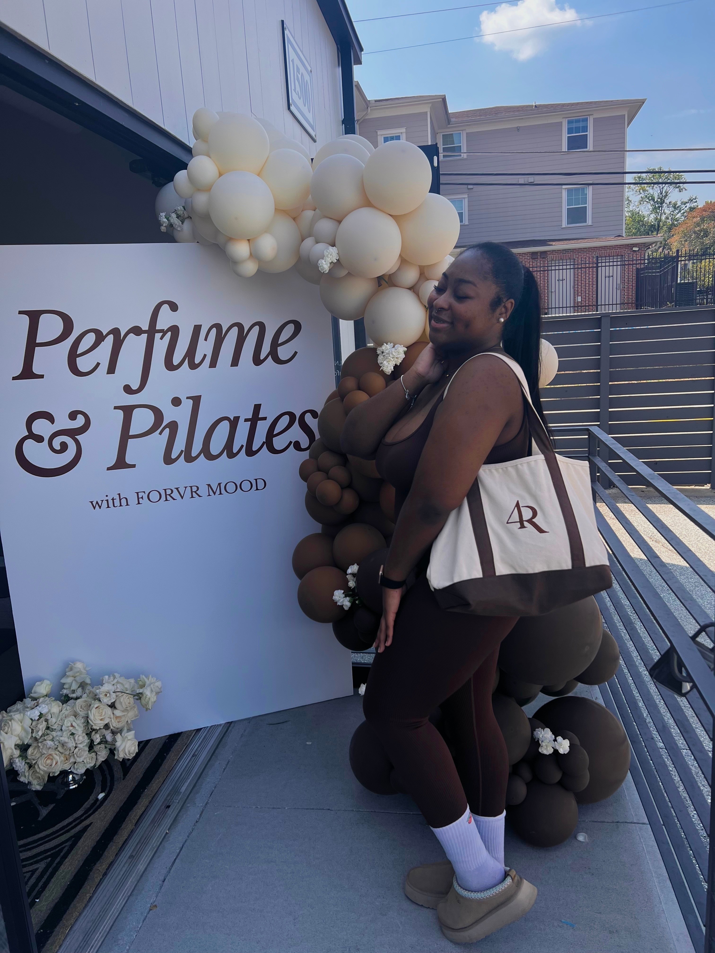 Smiling woman holding a bag, standing next to a perfume and Pilates event sign with balloon decorations outside.