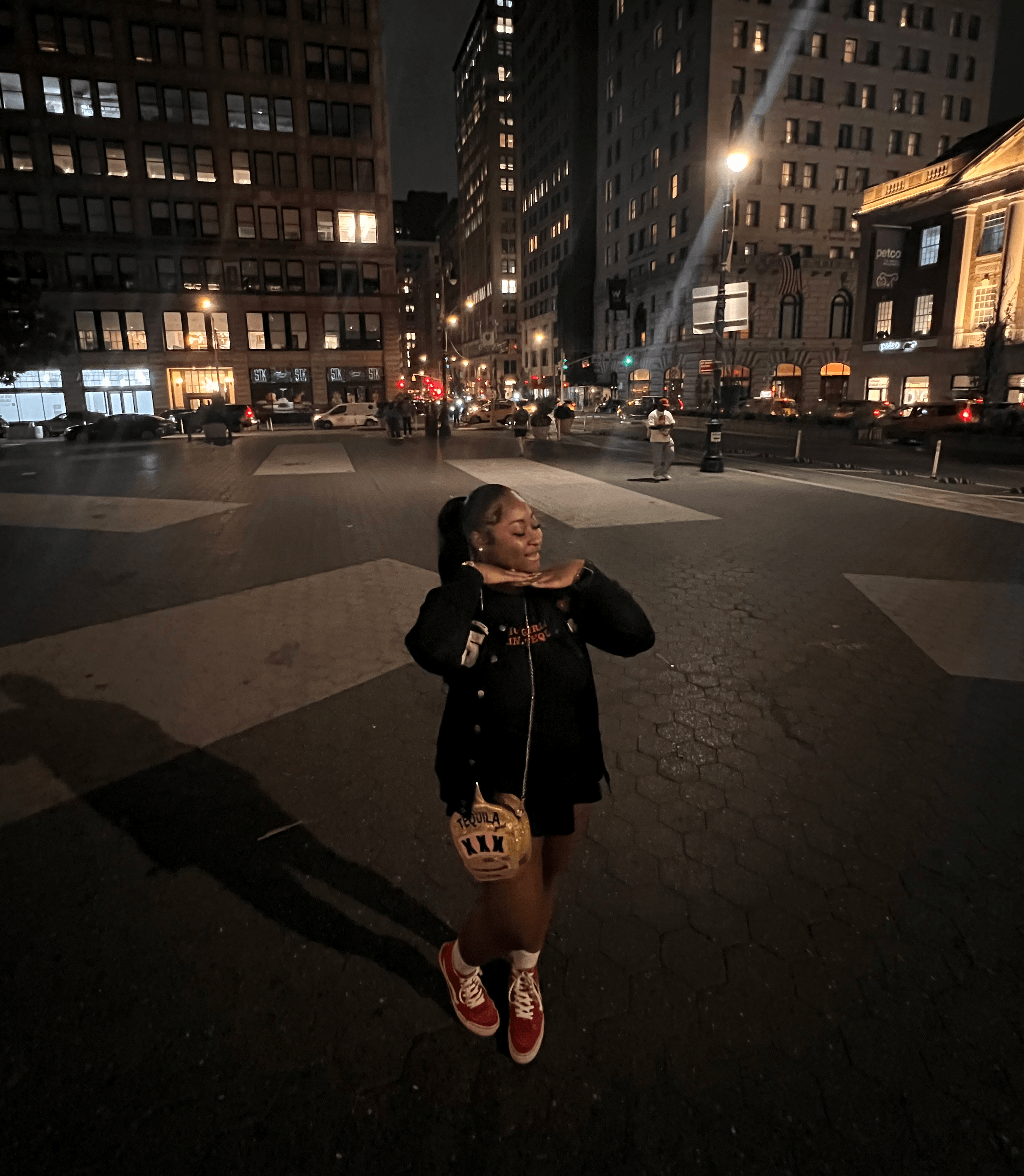 Young girl standing in a city street at night, surrounded by tall buildings with lit windows, wearing a black jacket and red shoes.