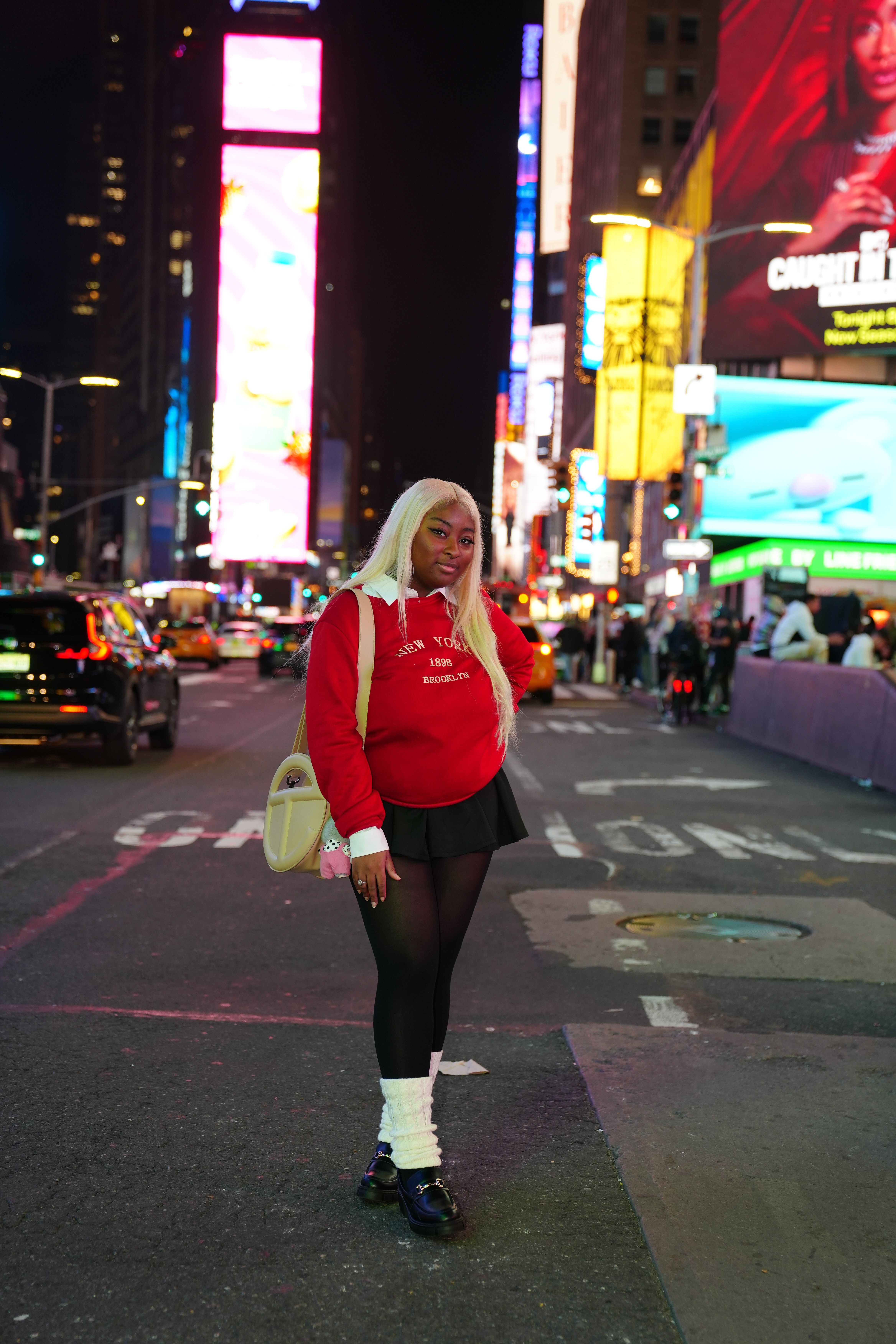 Person with blonde hair, red sweatshirt, black shorts, and white socks standing on city street at night with bright billboards.