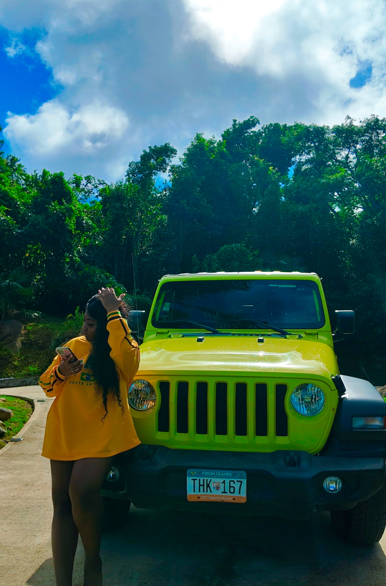 Person in yellow jacket standing in front of a bright green vehicle outdoors with trees and cloudy sky behind.
