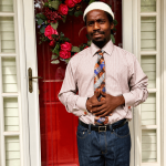 Man standing in front of a door with a red floral wreath, wearing a white hat, striped shirt, and tie.