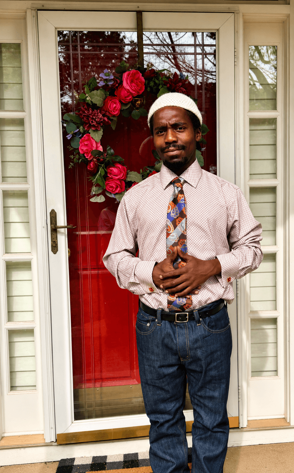 Man standing in front of a door with a red floral wreath, wearing a white hat, striped shirt, and tie.