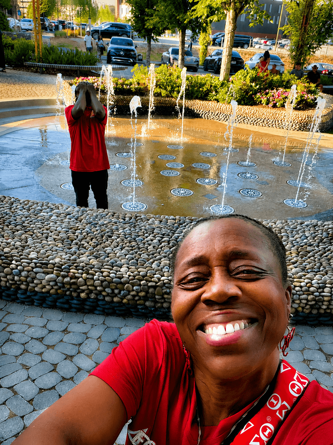 Smiling woman in red shirt taking selfie near a fountain with a person in red shirt in the background, trees, and parked cars.