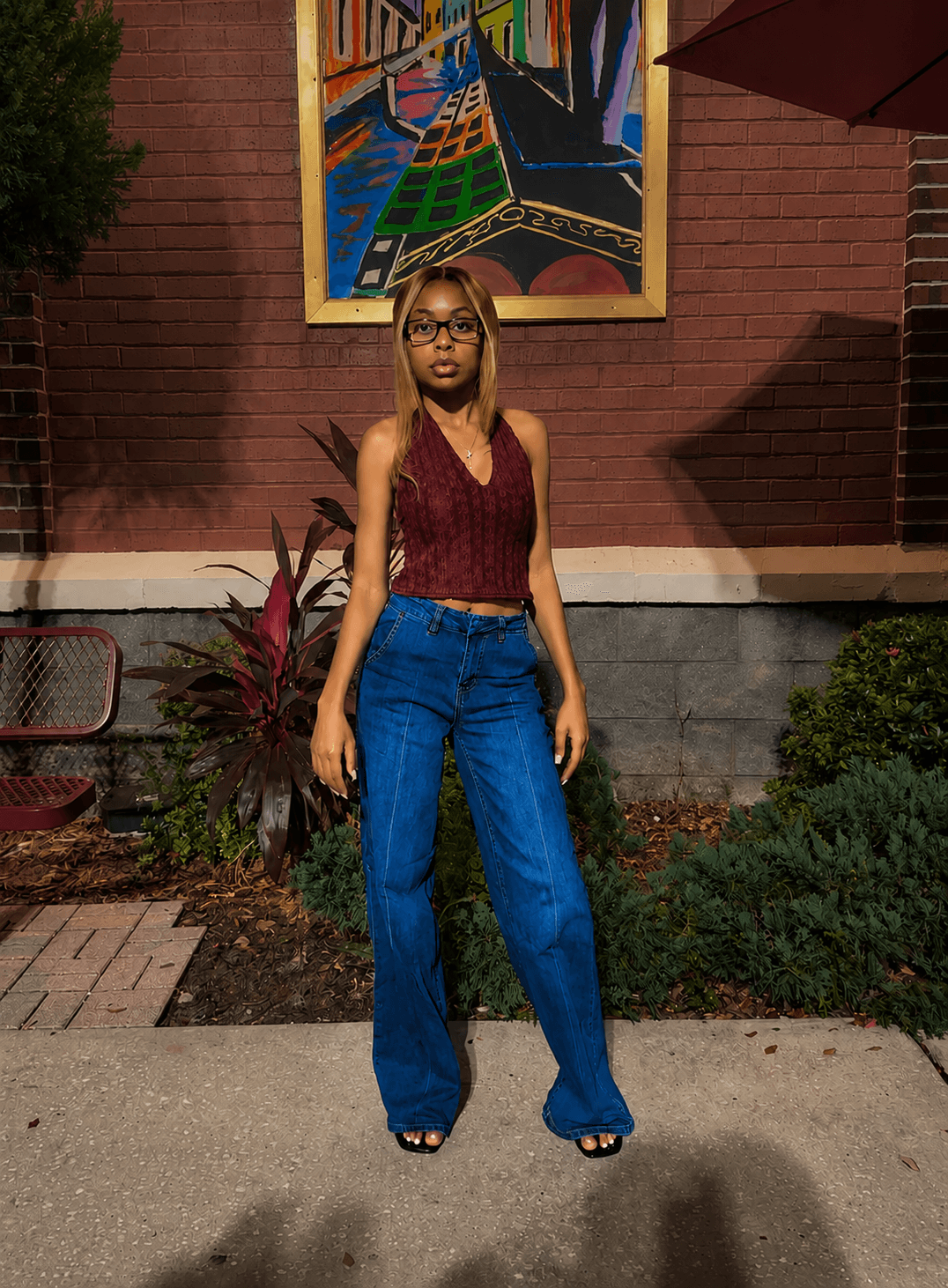 Young woman with glasses and long hair stands outdoors in front of a brick wall with colorful artwork, wearing a sleeveless top and jeans.