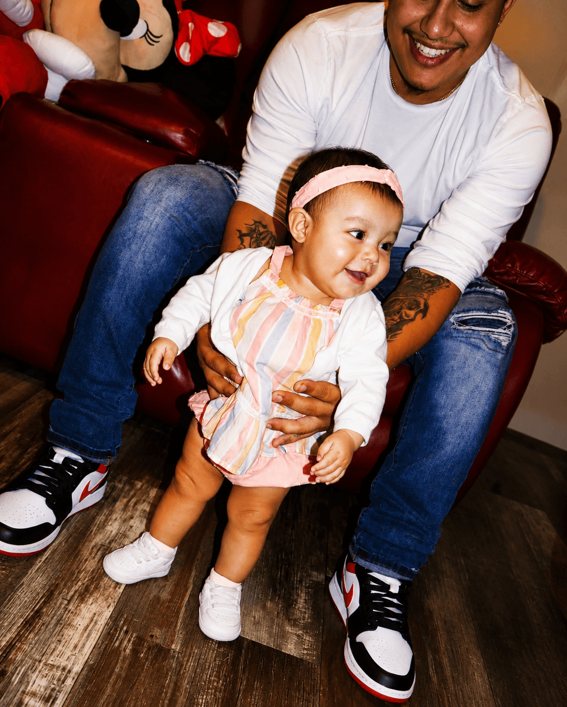 Adult smiling with a young girl, sitting on a red couch, with Mickey Mouse plush toys nearby, wooden floor visible.