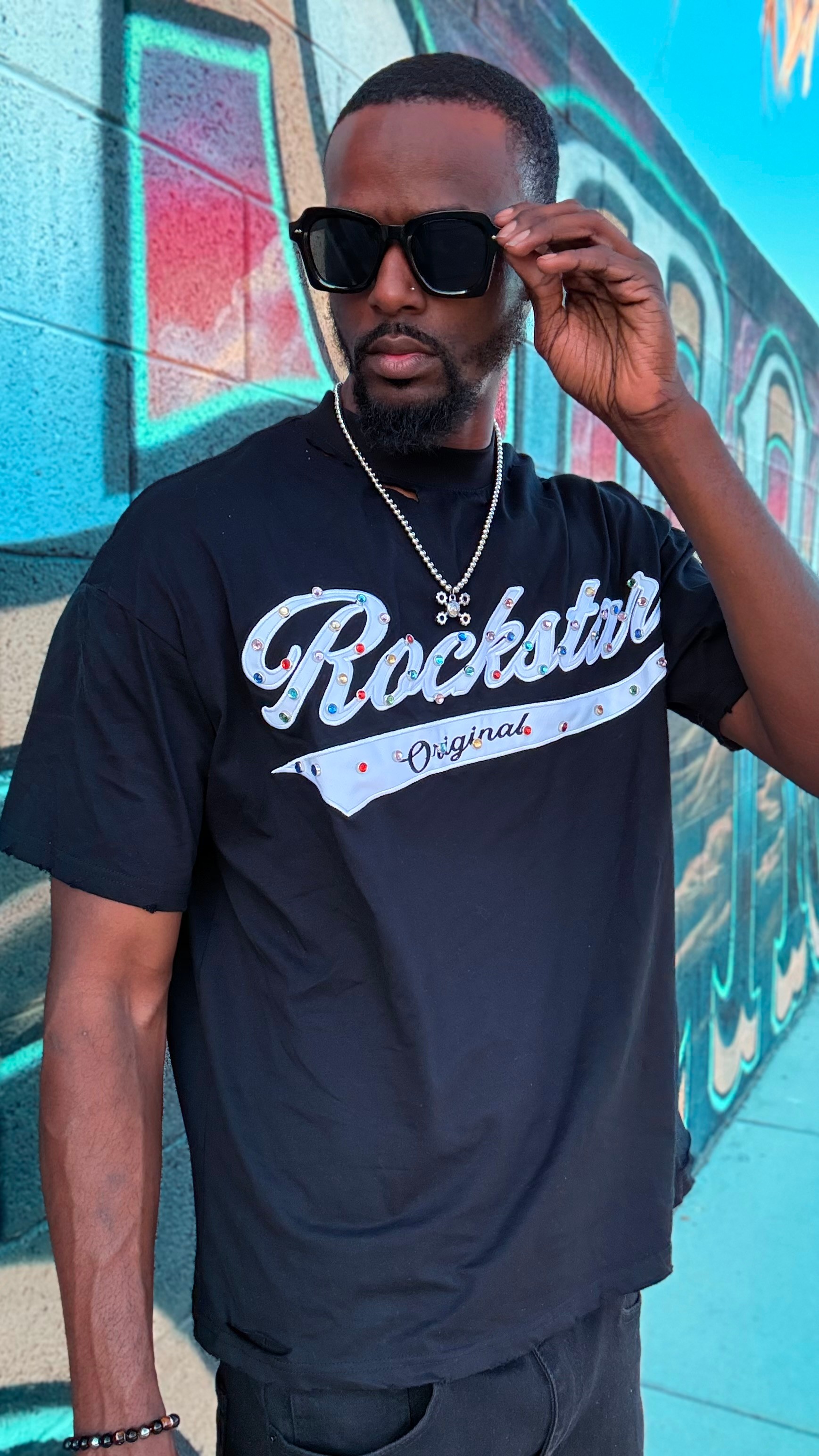 Man wearing sunglasses and a black T-shirt with 'Rockstar' text, standing in front of a colorful graffiti wall.