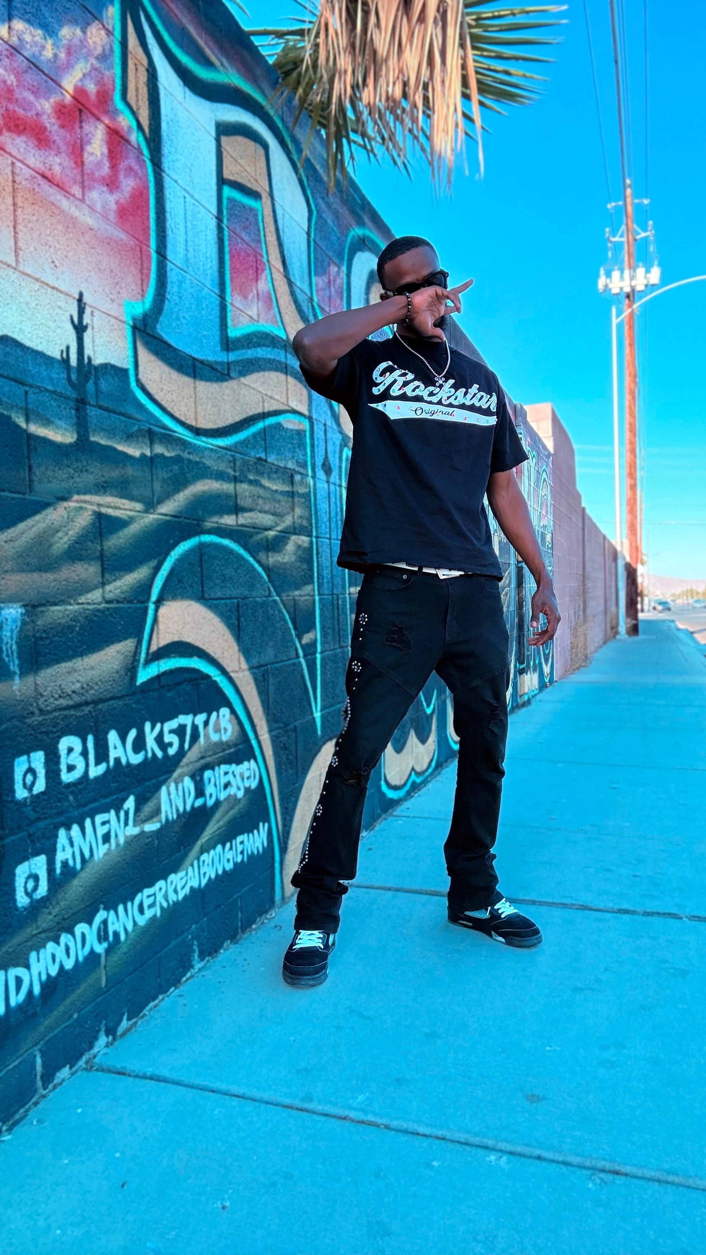 Person standing on sidewalk in front of graffiti wall, wearing sunglasses, black t-shirt, and pants, pointing at camera.