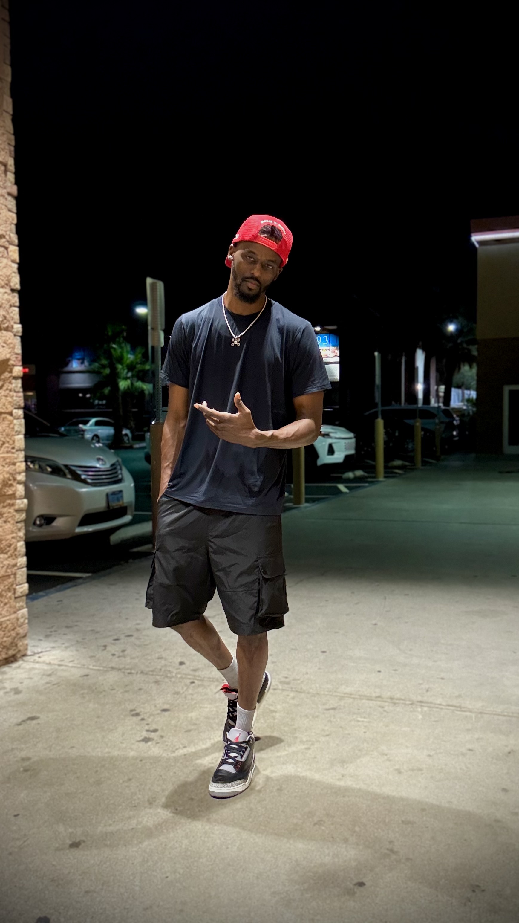 Young man standing outdoors at night, wearing a red cap, black t-shirt, shorts, and sneakers, making a hand gesture.