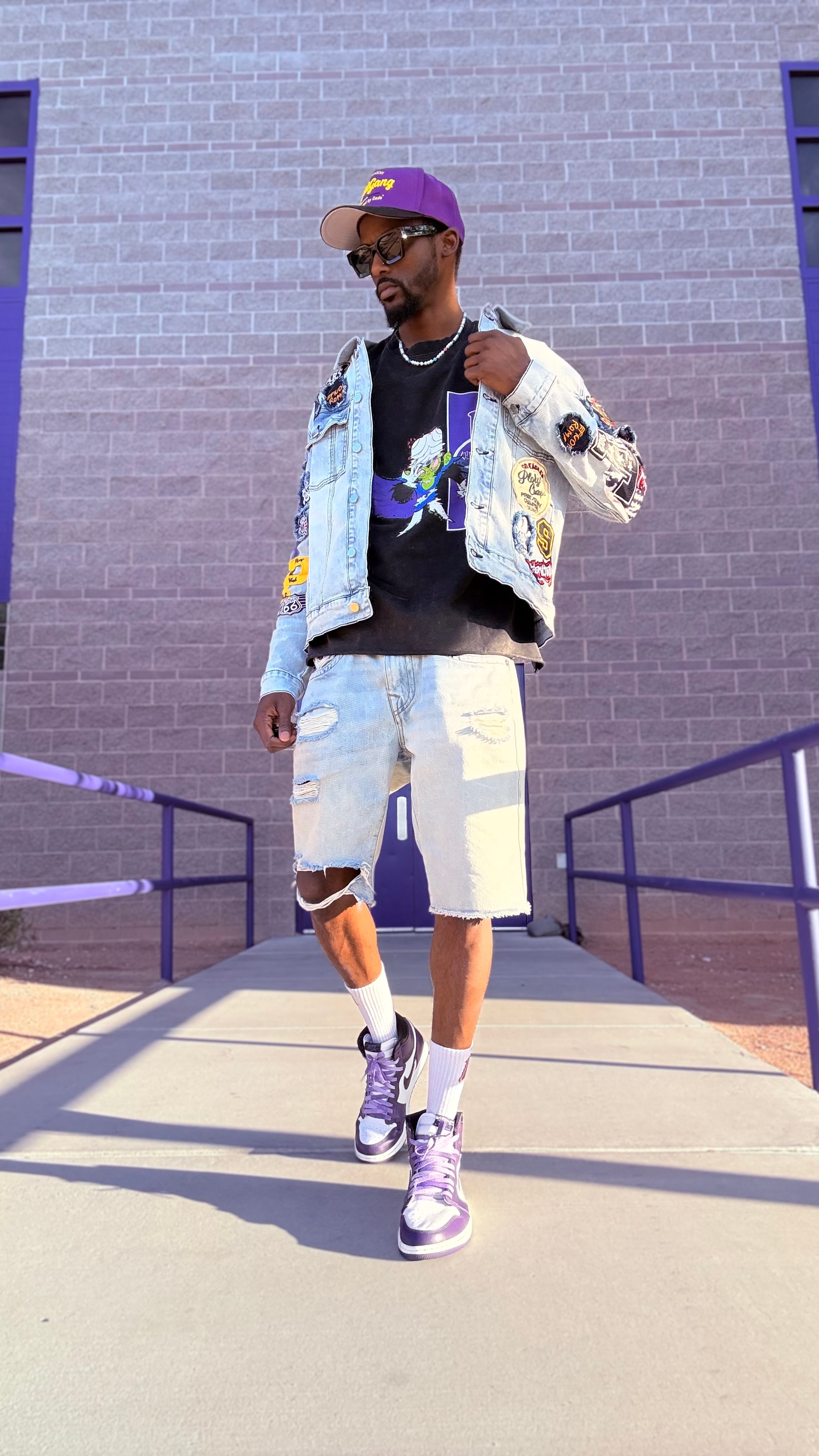Man in colorful jacket, black shirt, ripped shorts, purple cap, and sneakers walking outdoors against purple wall