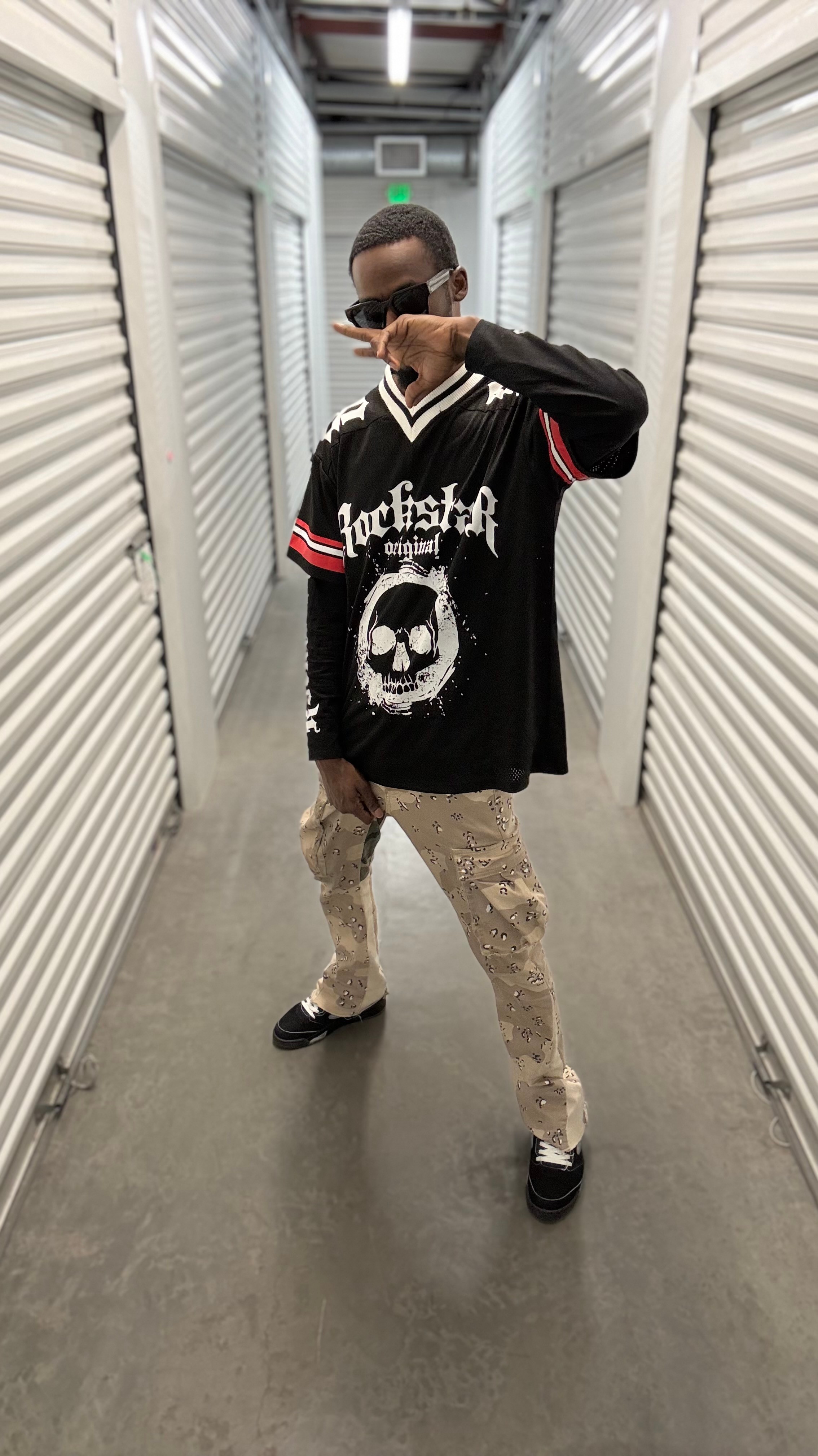 Person taking a mirror selfie in a storage unit hallway, wearing a black graphic sweatshirt, beige pants, and sunglasses.
