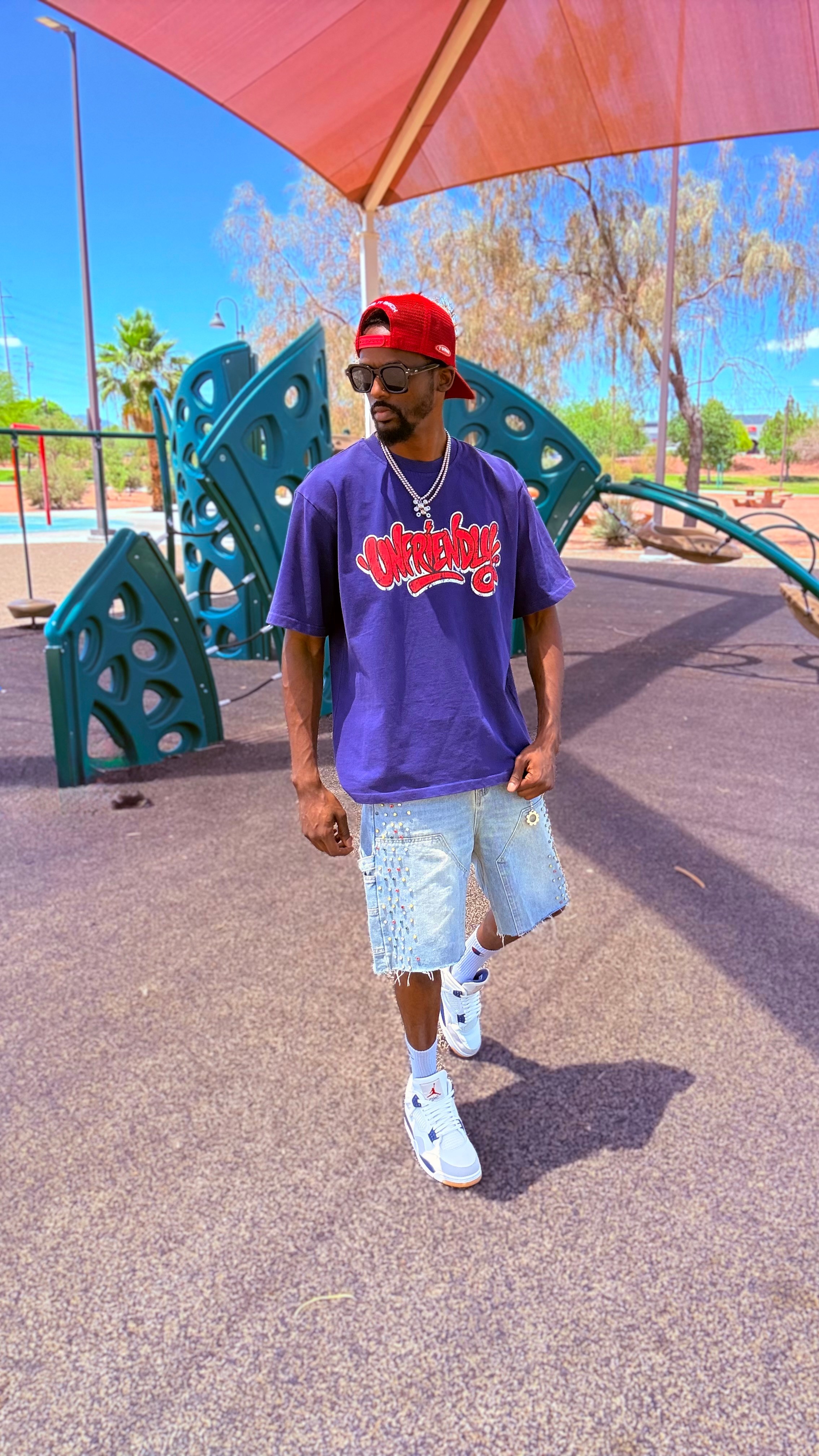 Person walking in a park with playground equipment, wearing a purple shirt, denim shorts, white shoes, and a red cap.