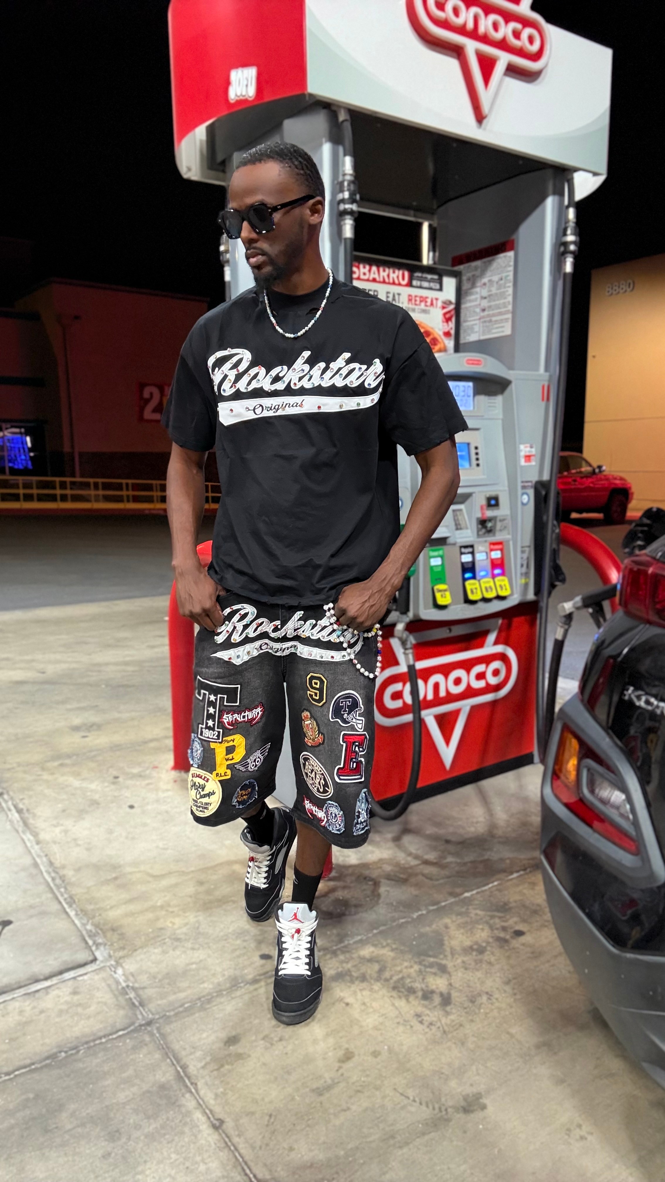 Man in sunglasses and black t-shirt with 'Rockstar' logo at gas station, wearing patterned shorts and sneakers, standing near fuel pump.