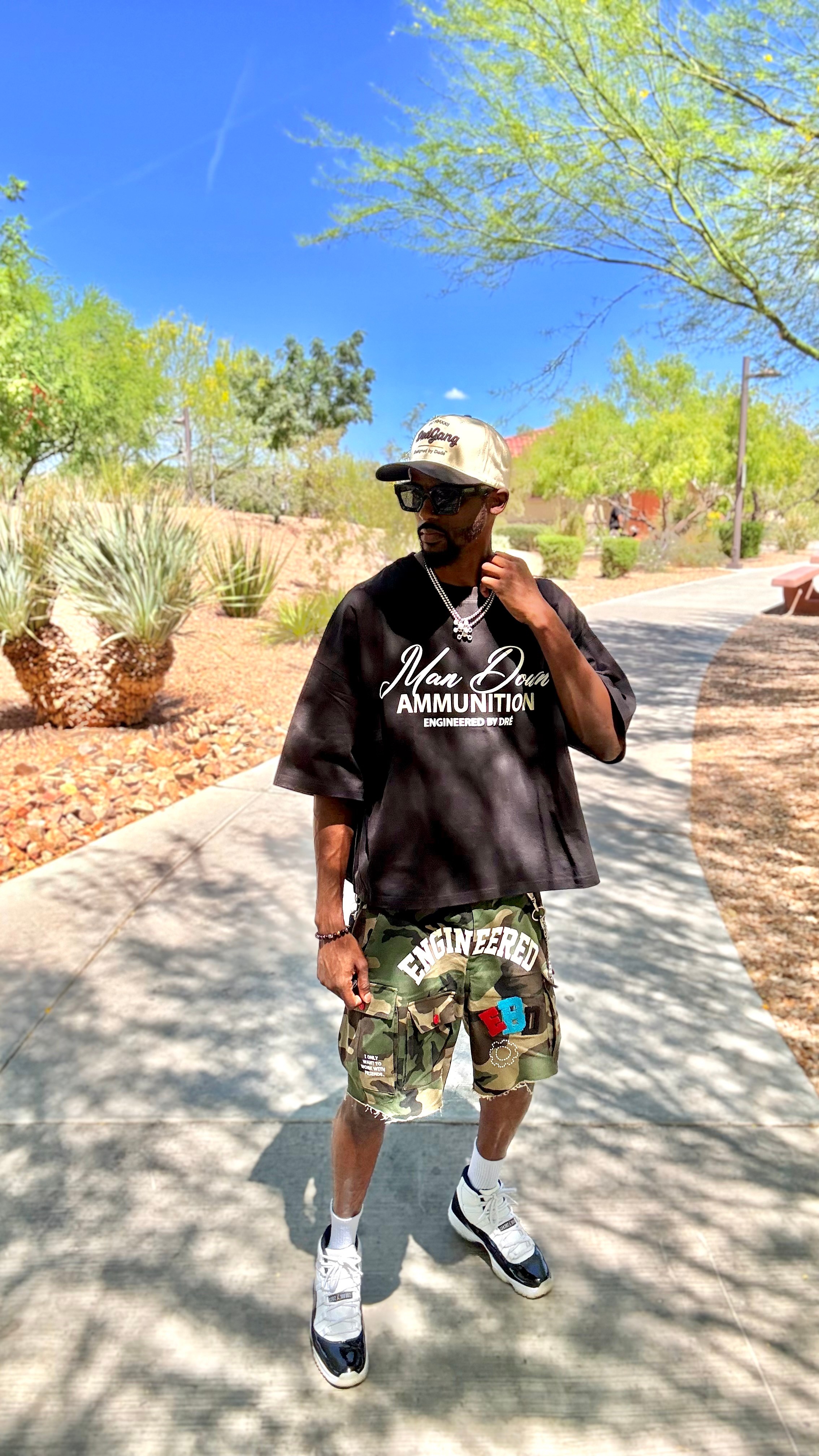 Person walking outdoors on a sunny day, wearing a black t-shirt, camouflage shorts, and a hat, with trees and blue sky in background.