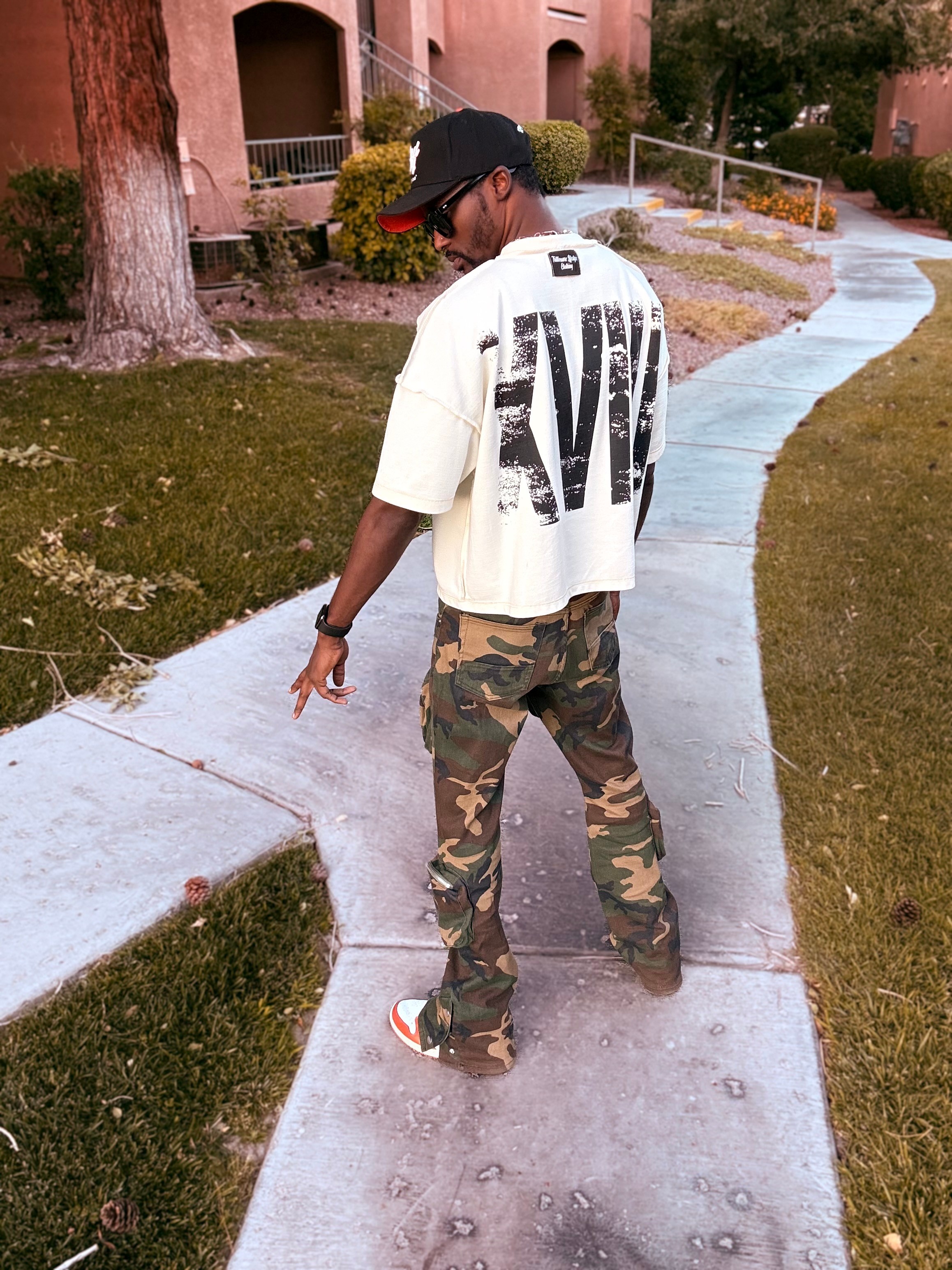 Person walking on a sidewalk in a residential area, wearing a white oversized T-shirt, camouflage pants, and a black cap.