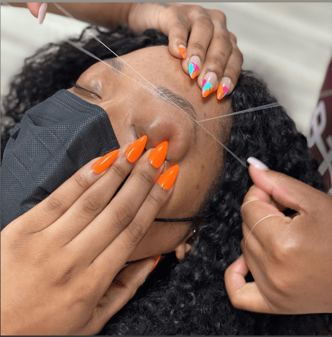 Person receiving eyelash extension treatment, with hands holding eye and eyelash tools, nails painted bright orange and multicolored. Word partially visible in background.