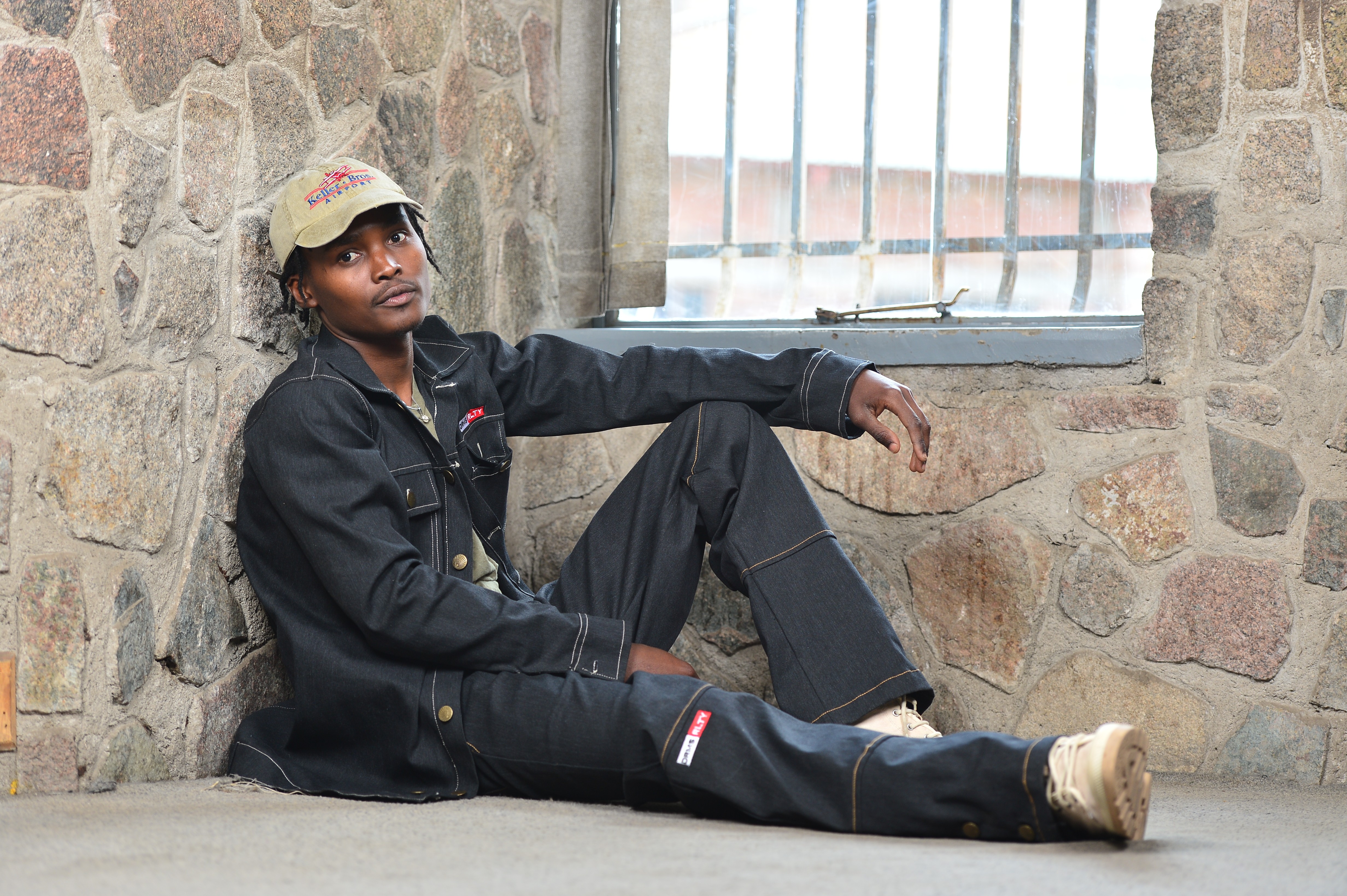 Young person sitting on the floor against a stone wall near a window, wearing a black jacket and pants, beige cap, and beige shoes.