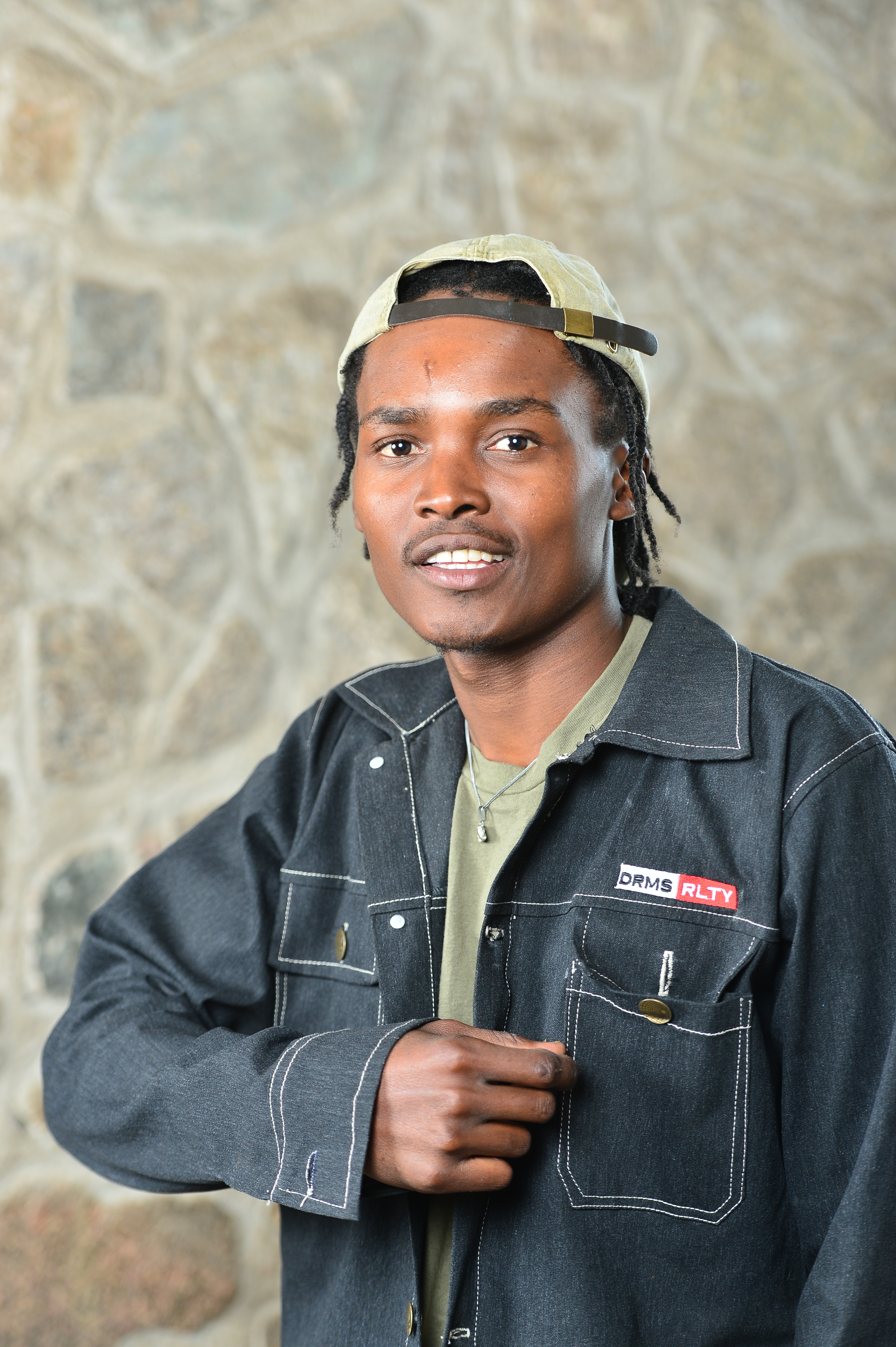 Young man wearing a black denim jacket and a cap backwards, standing outdoors with a stone wall background.
