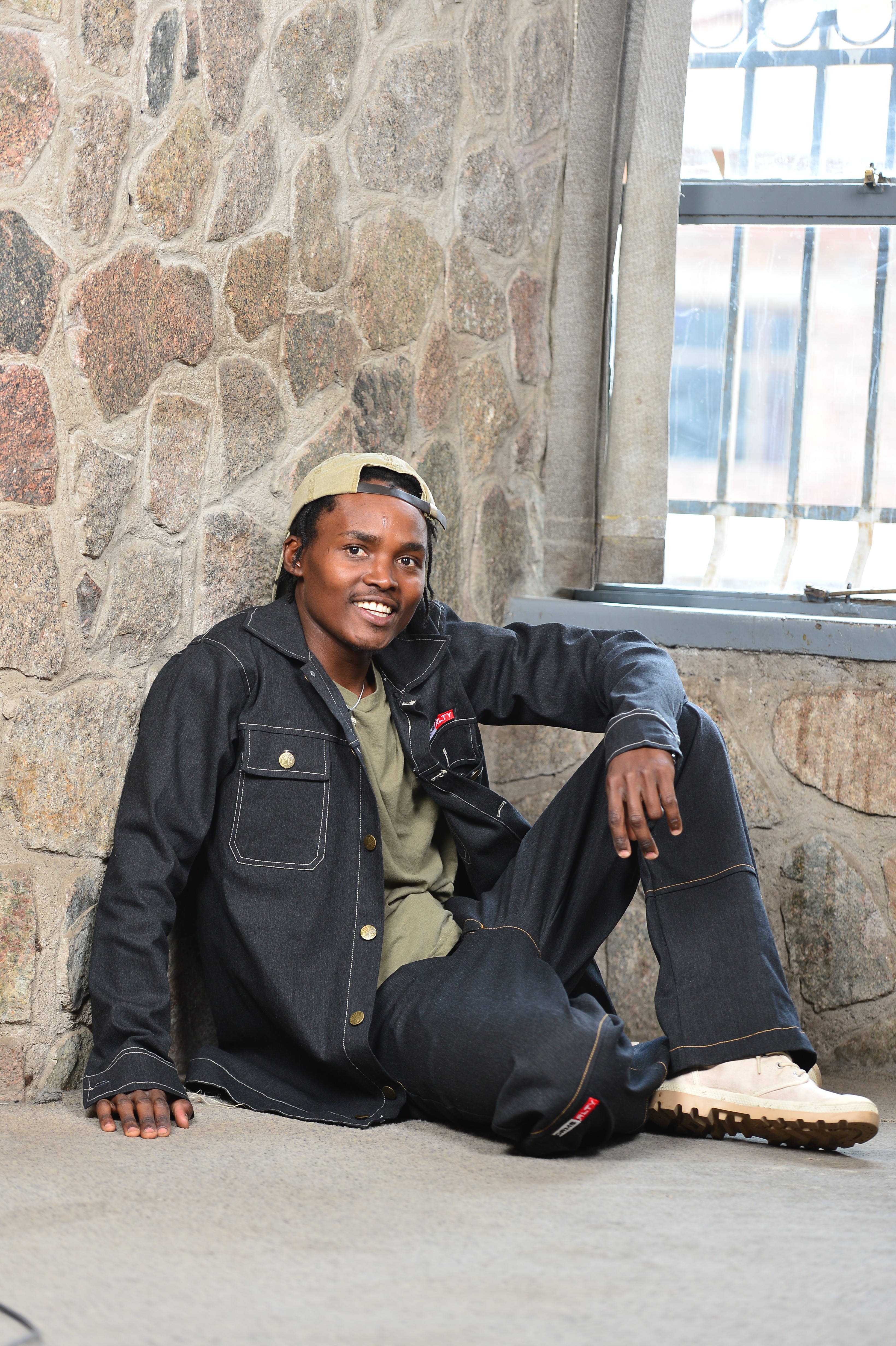 Person sitting on the ground against a stone wall near a window, smiling, wearing a black jacket and beige shoes.