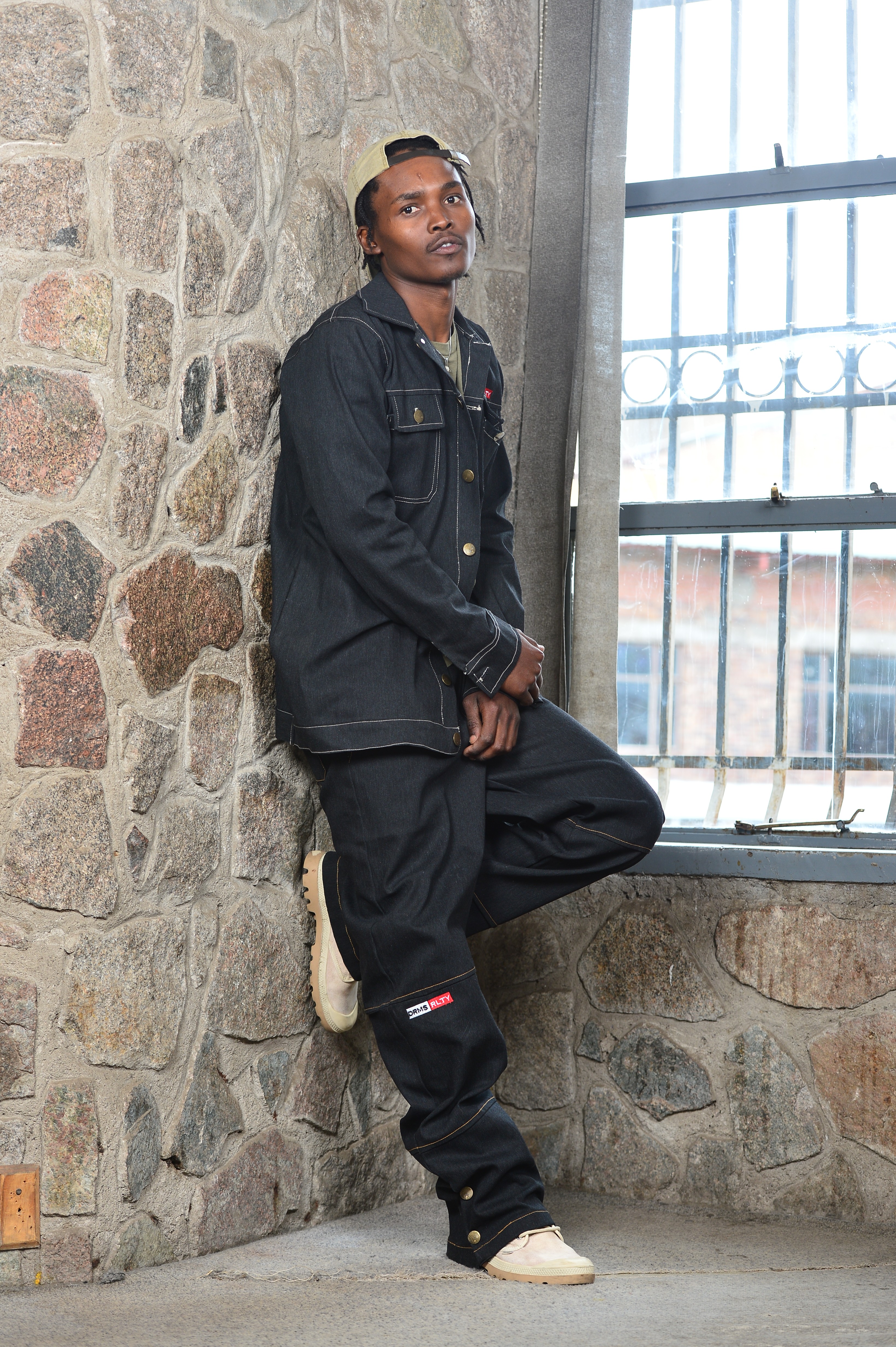 Young man leaning against a stone wall near a large window, wearing a black denim jacket and pants, holding a phone.