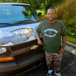 Man standing next to a black vehicle with a logo, outdoors with trees and sunlight, wearing a green Operation Defrost T-shirt and camouflage pants.