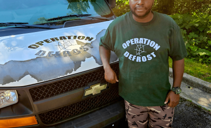 Man standing next to a black vehicle with a logo, outdoors with trees and sunlight, wearing a green Operation Defrost T-shirt and camouflage pants.