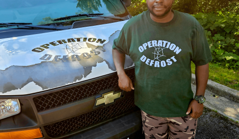 Man standing next to a black vehicle with a logo, outdoors with trees and sunlight, wearing a green Operation Defrost T-shirt and camouflage pants.