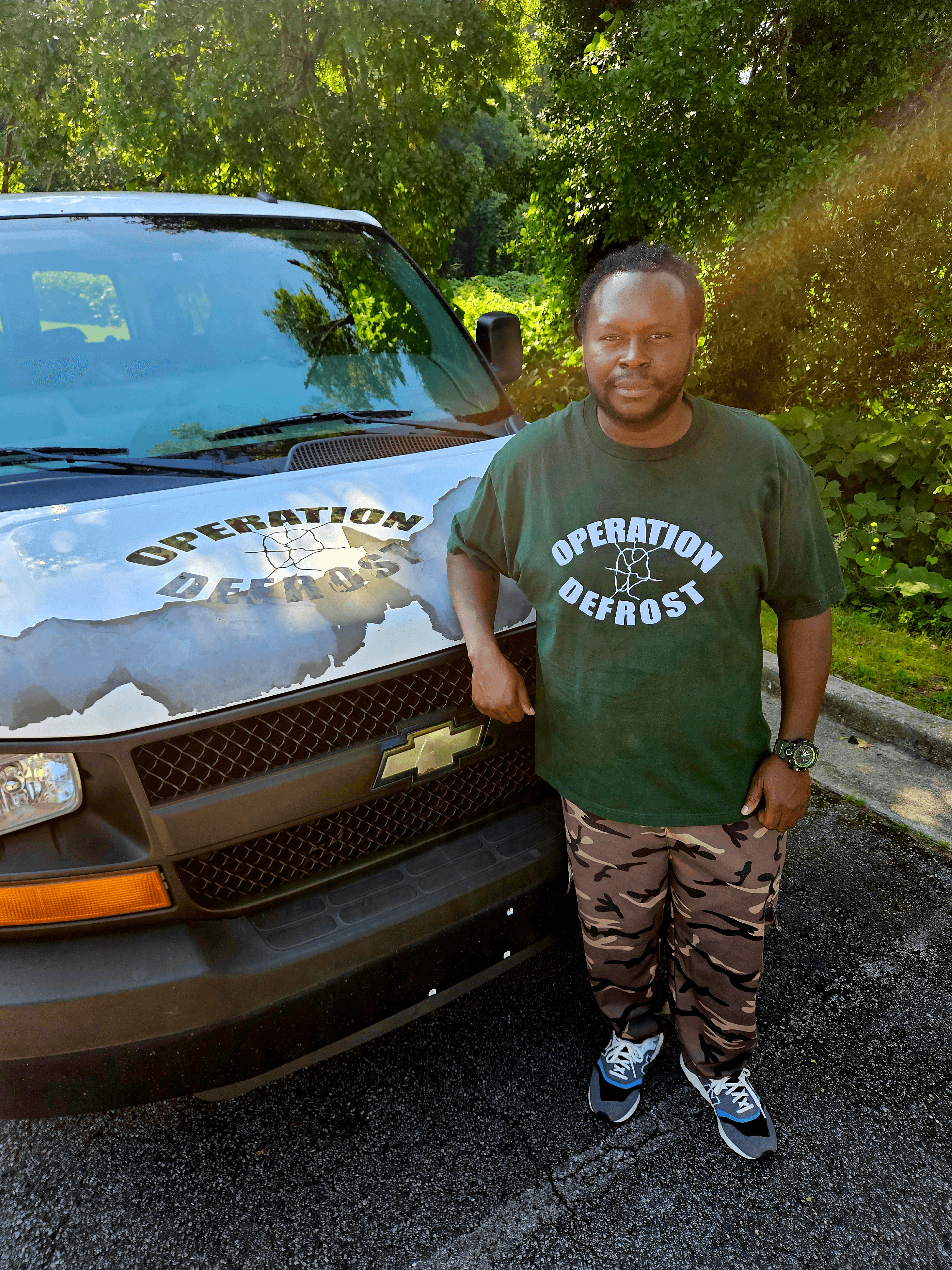 Man standing next to a black vehicle with a logo, outdoors with trees and sunlight, wearing a green Operation Defrost T-shirt and camouflage pants.